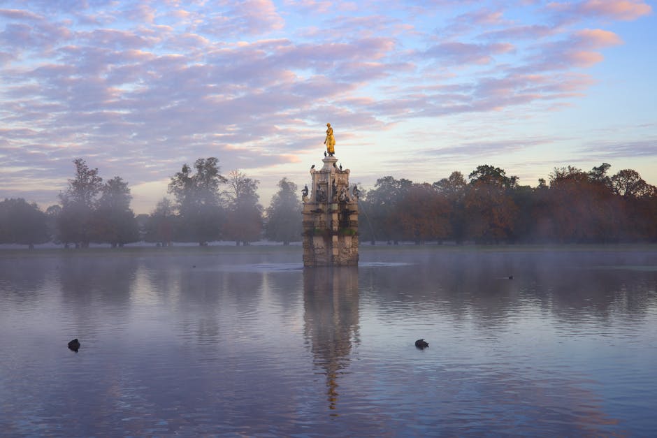 A serene lake scene during early morning with calm, reflective waters and a light mist hovering over the surface. In the center of the lake stands an ornate stone monument topped with a gilded statue,